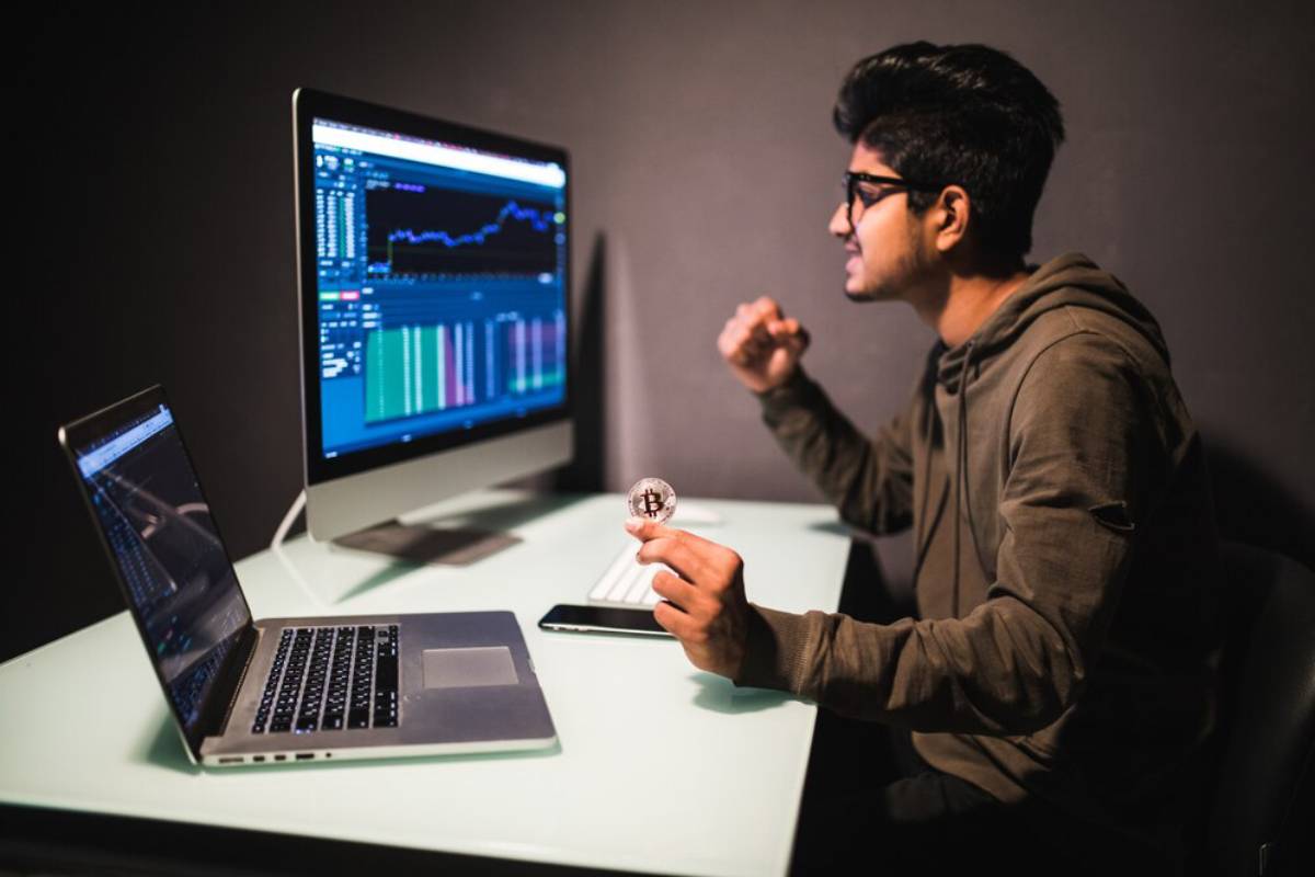 Young man examines Bitcoin while working with multiple computers displaying financial data.