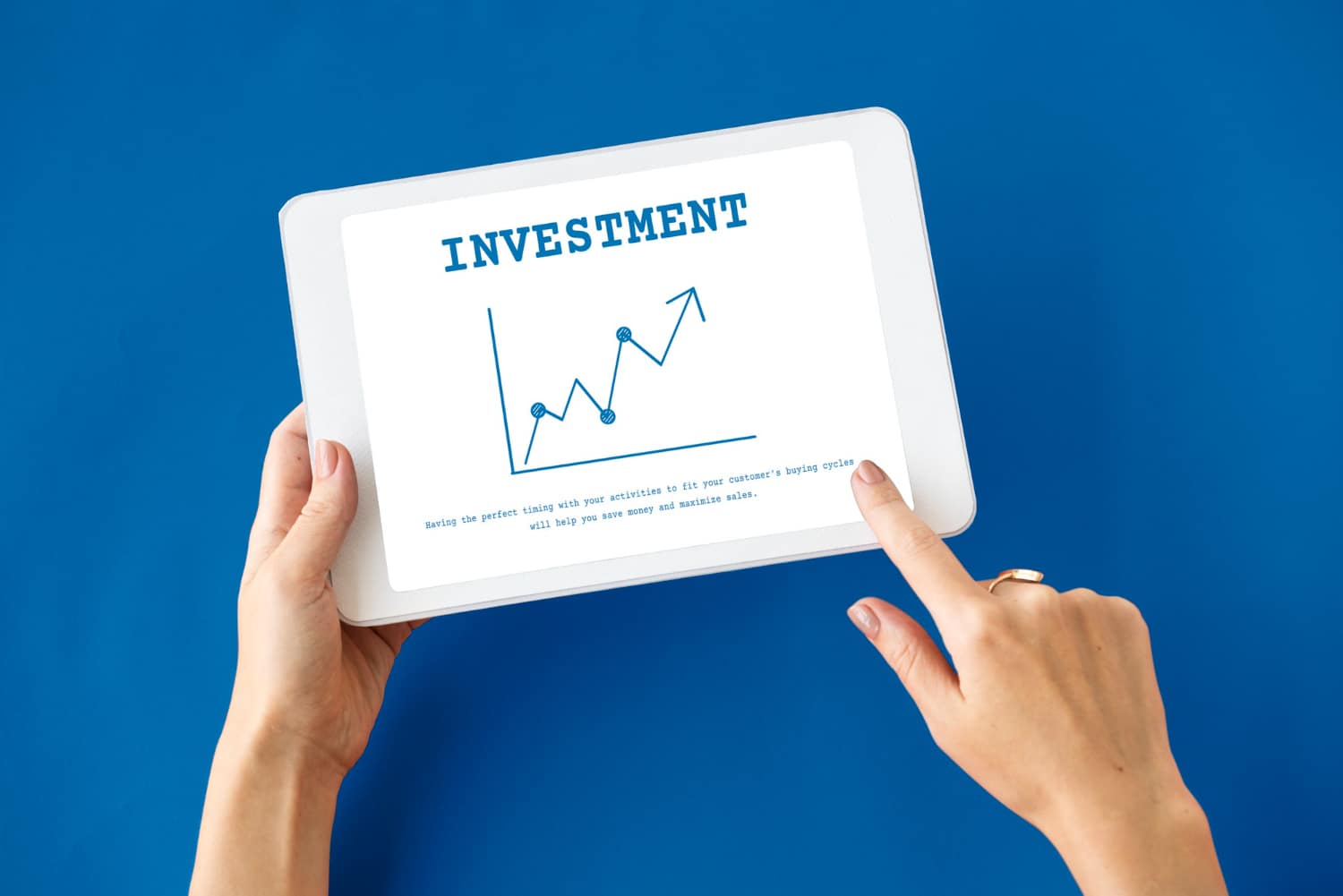 Hands holding a tablet displaying a graph labeled INVESTMENT with ascending trend lines, against a blue background.