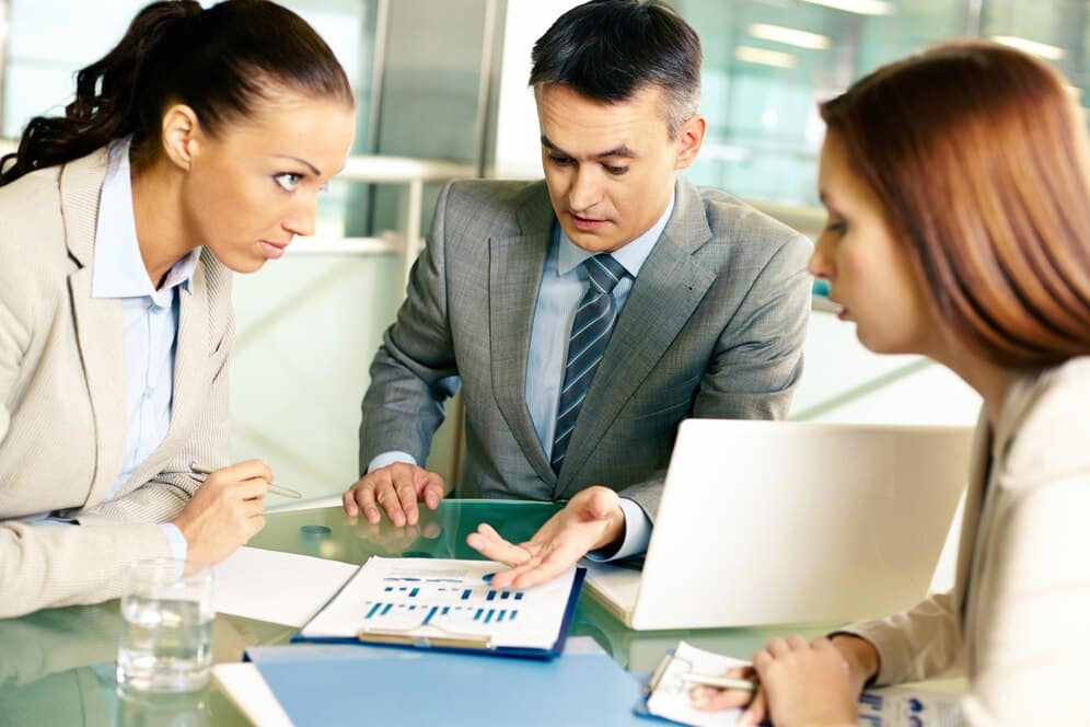 Three professionals discussing graphs and documents at a glass table in a modern office setting.
