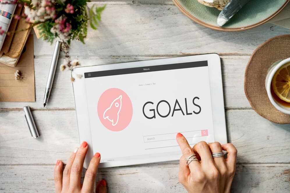 Hands on a tablet displaying GOALS on screen, with a tea cup and stationary on a wooden table.