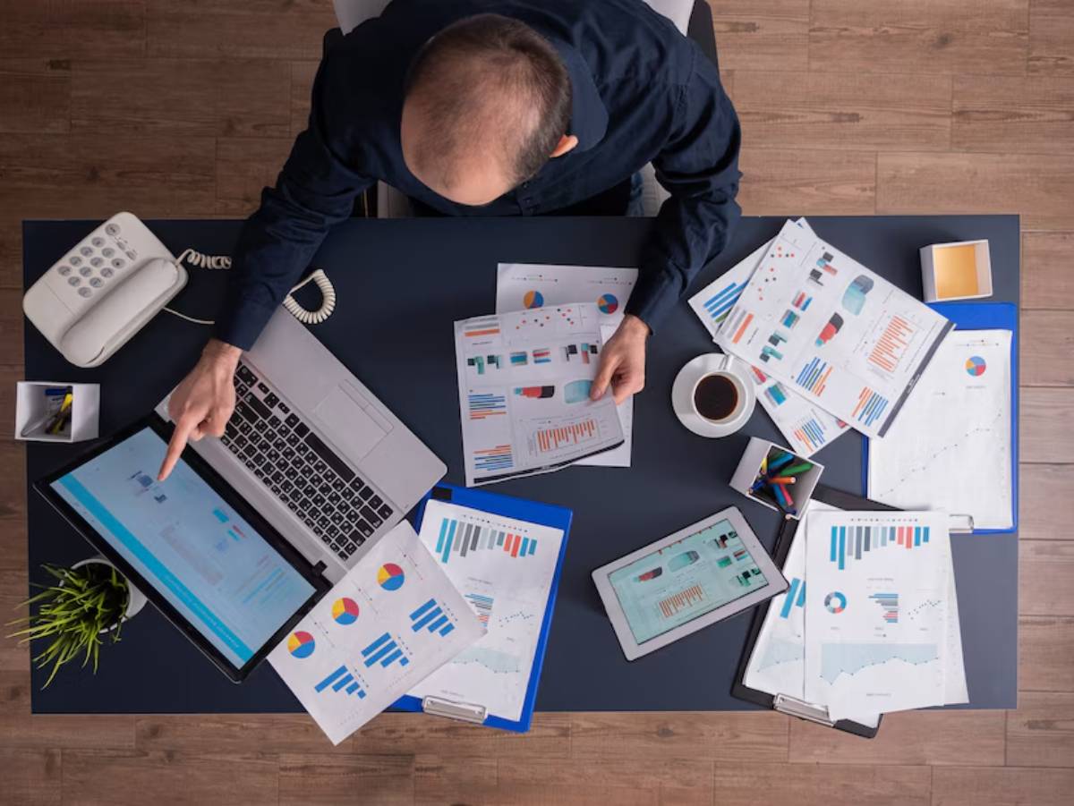 Top-down view of a man analyzing graphs on multiple documents with a laptop and mobile phone on a desk.