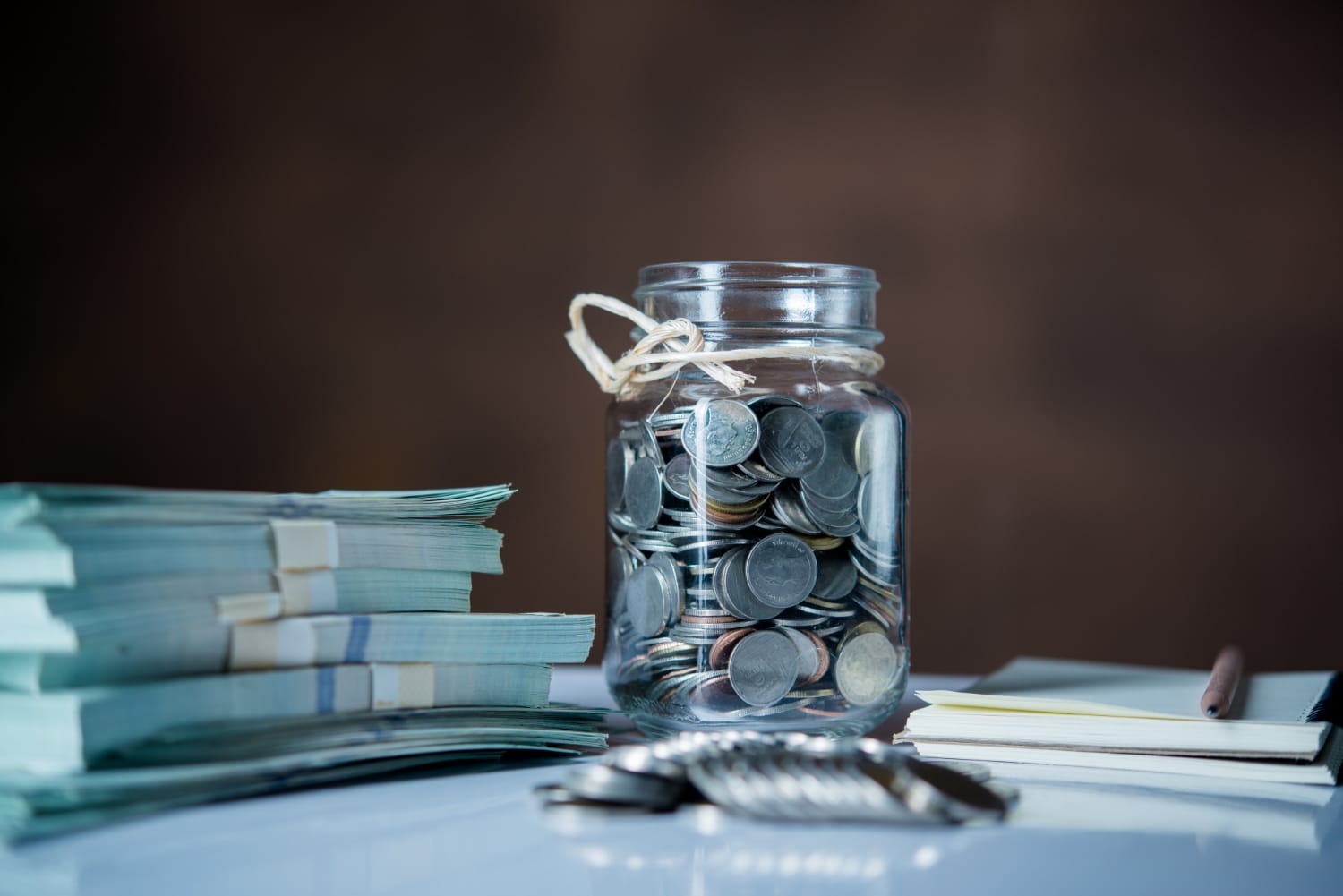 A glass jar filled with coins, tied with twine, beside stacks of banknotes and a pen on a table.