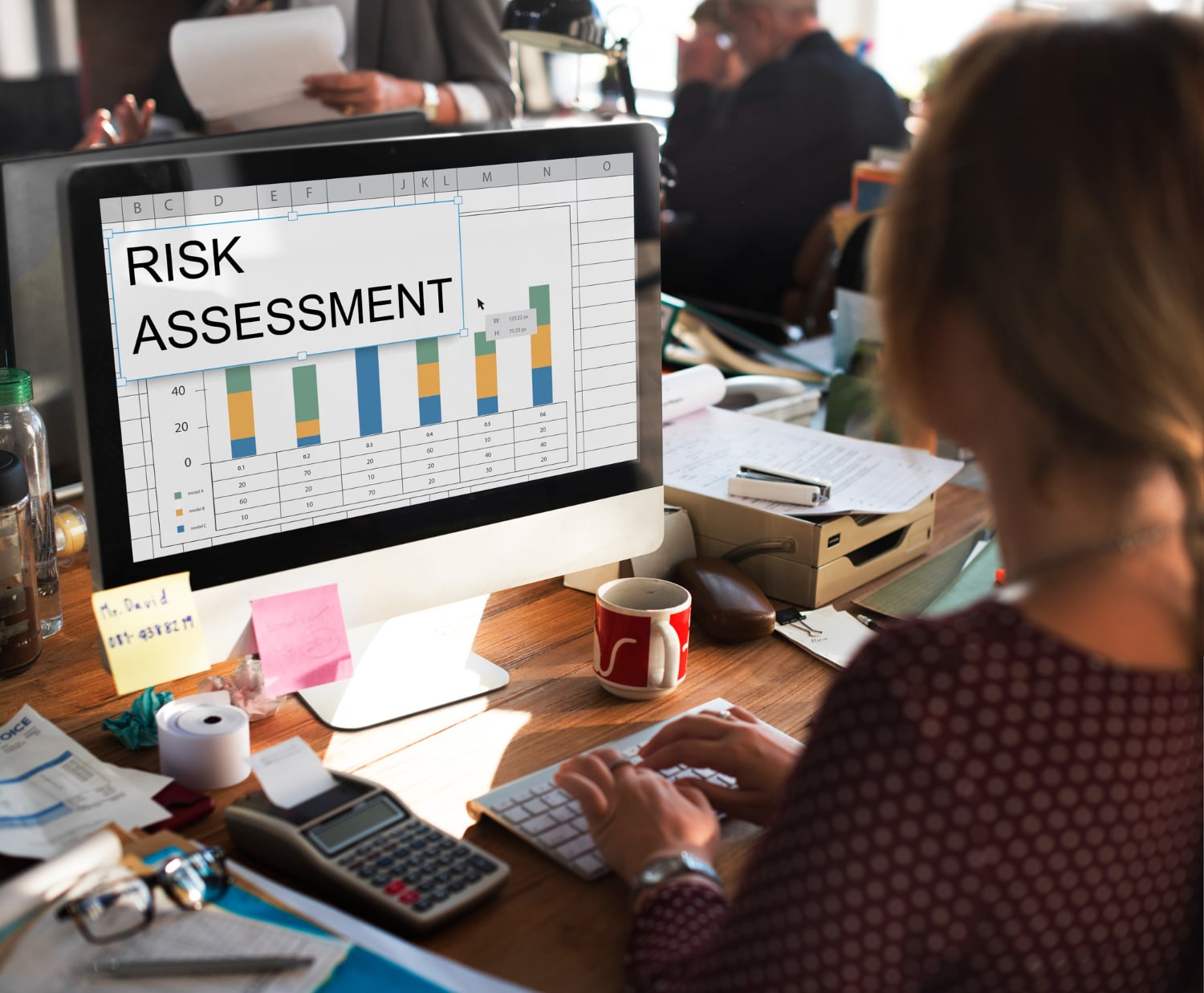 Woman analyzing a Risk Assessment bar chart on her computer in a busy office environment.