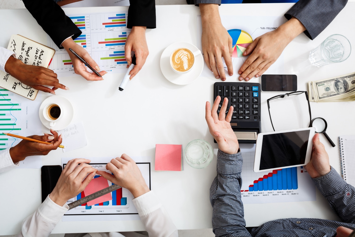 Top view of a business meeting with hands pointing at financial charts and electronic devices on a table.