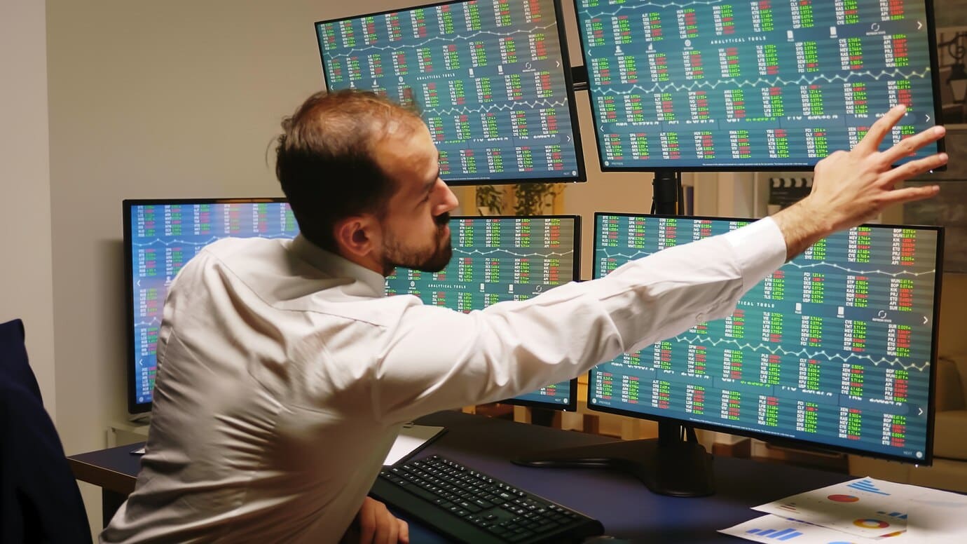 Man in a white shirt reaches towards multiple screens displaying financial data in an office environment.