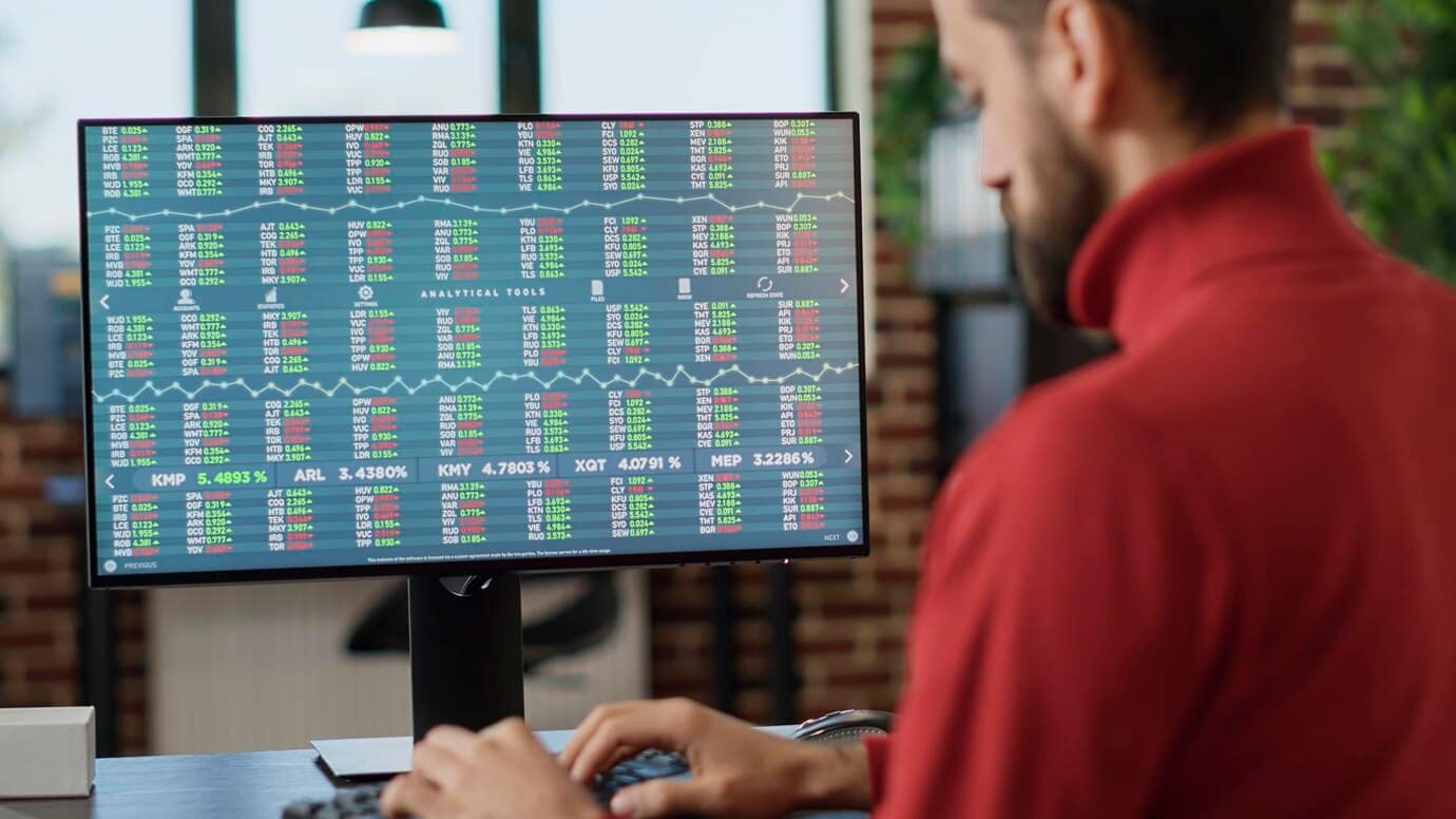 Man in red shirt analyzing data on a large computer screen displaying colorful financial charts.