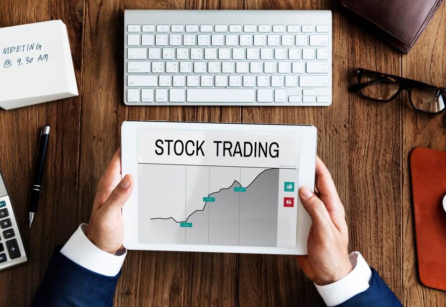 Person viewing stock trading graph on a tablet, with office items like keyboard and glasses nearby.