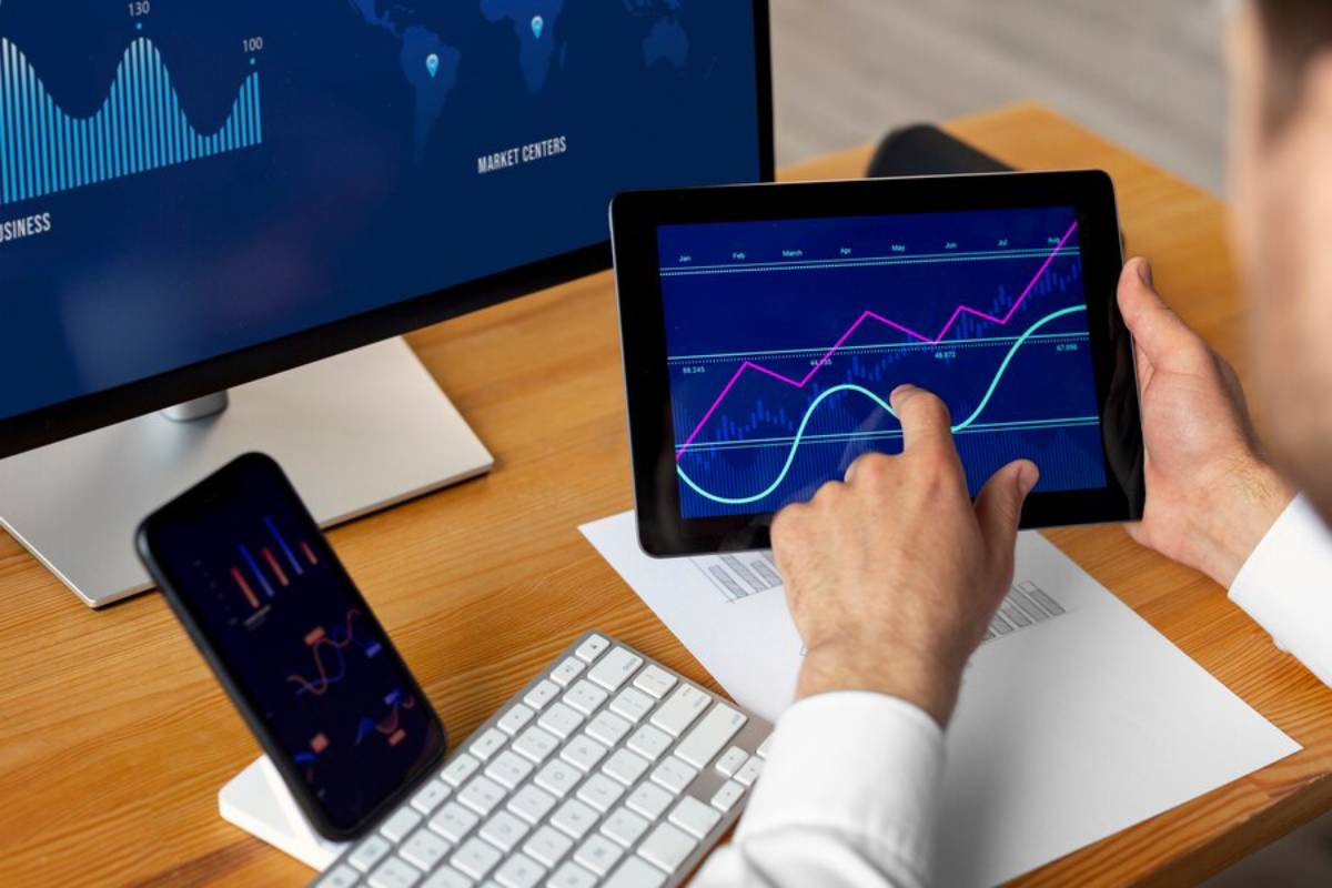 Businessman analyzing financial graphs on a tablet, with additional data displayed on a monitor and smartphone.