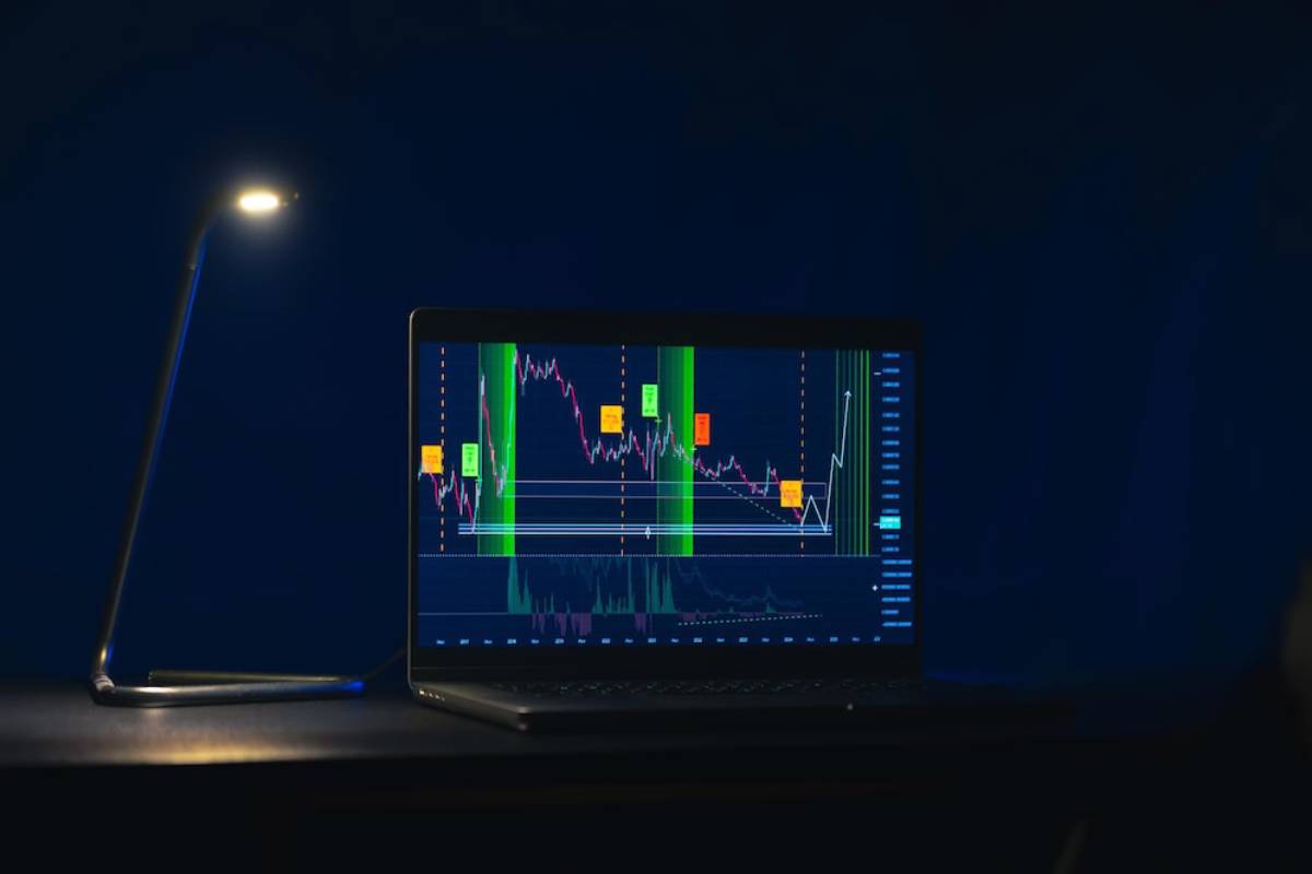Laptop displaying stock market charts on screen under a desk lamp, in a dark room.