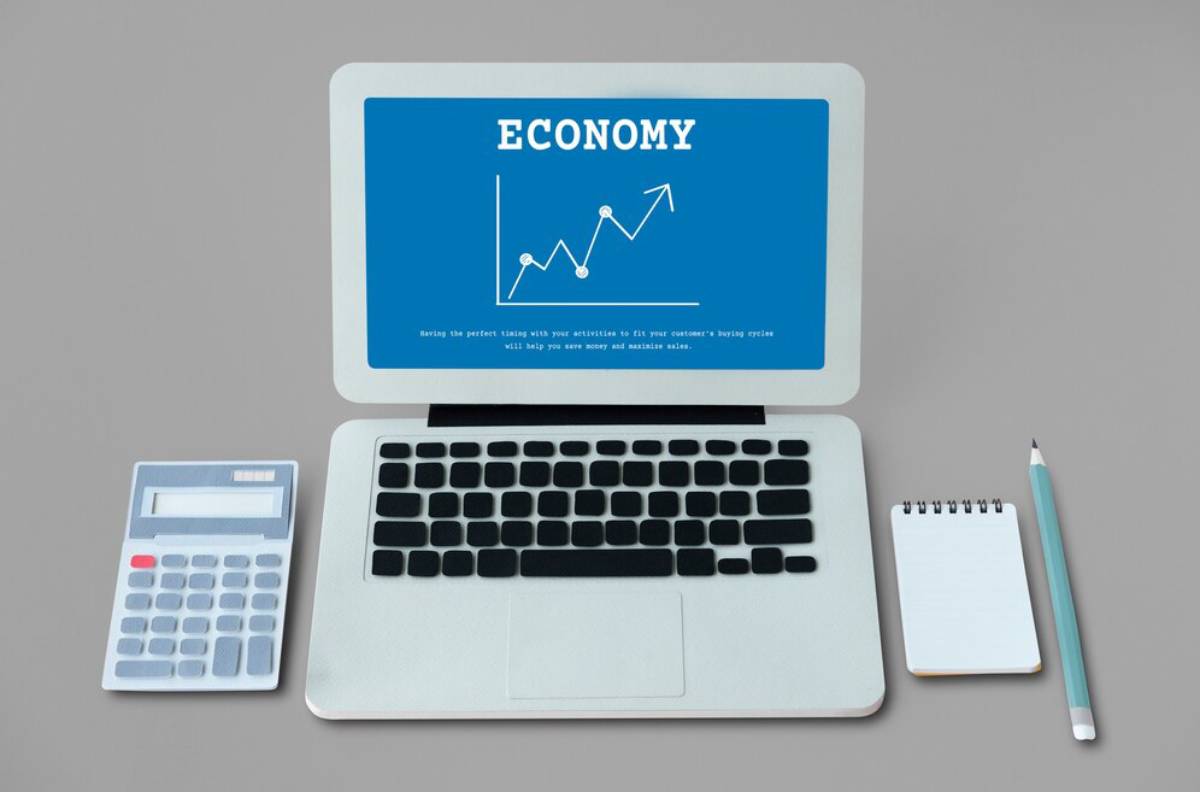A laptop displaying a presentation on economy with a graph, alongside a calculator, notebook, and pencil on a desk.