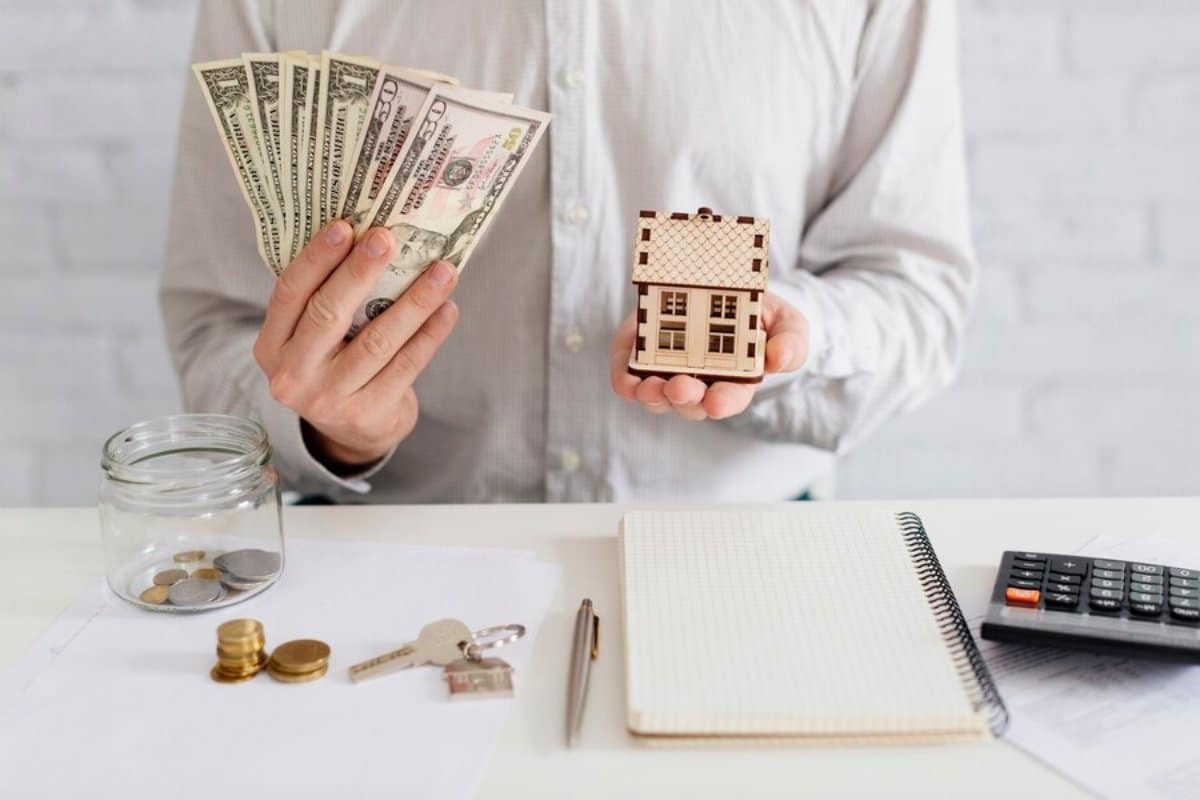 Person holding cash and a model house, with financial items on a desk, symbolizing budgeting and real estate investment.