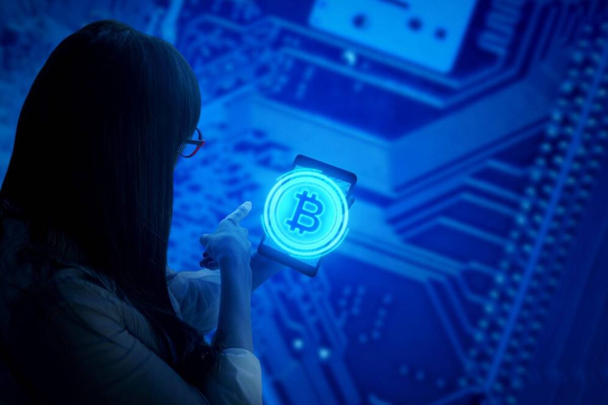 Woman in glasses interacting with a 3D hologram of a Bitcoin symbol, against a digital circuit board background.