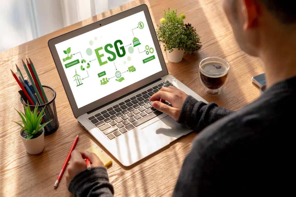 Person using a laptop displaying "ESG" environmental, social, and governance concepts, with office supplies and a cup of coffee on the desk.