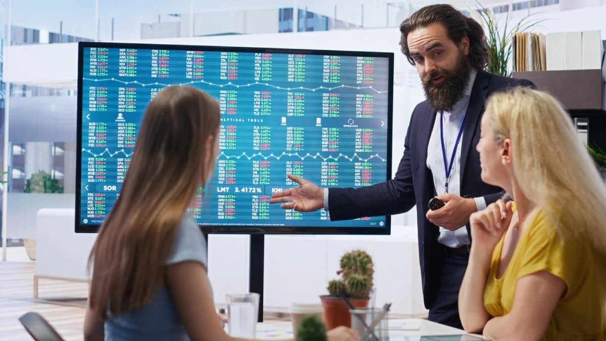 A bearded man explaining financial data on a large screen to two attentive women in an office.