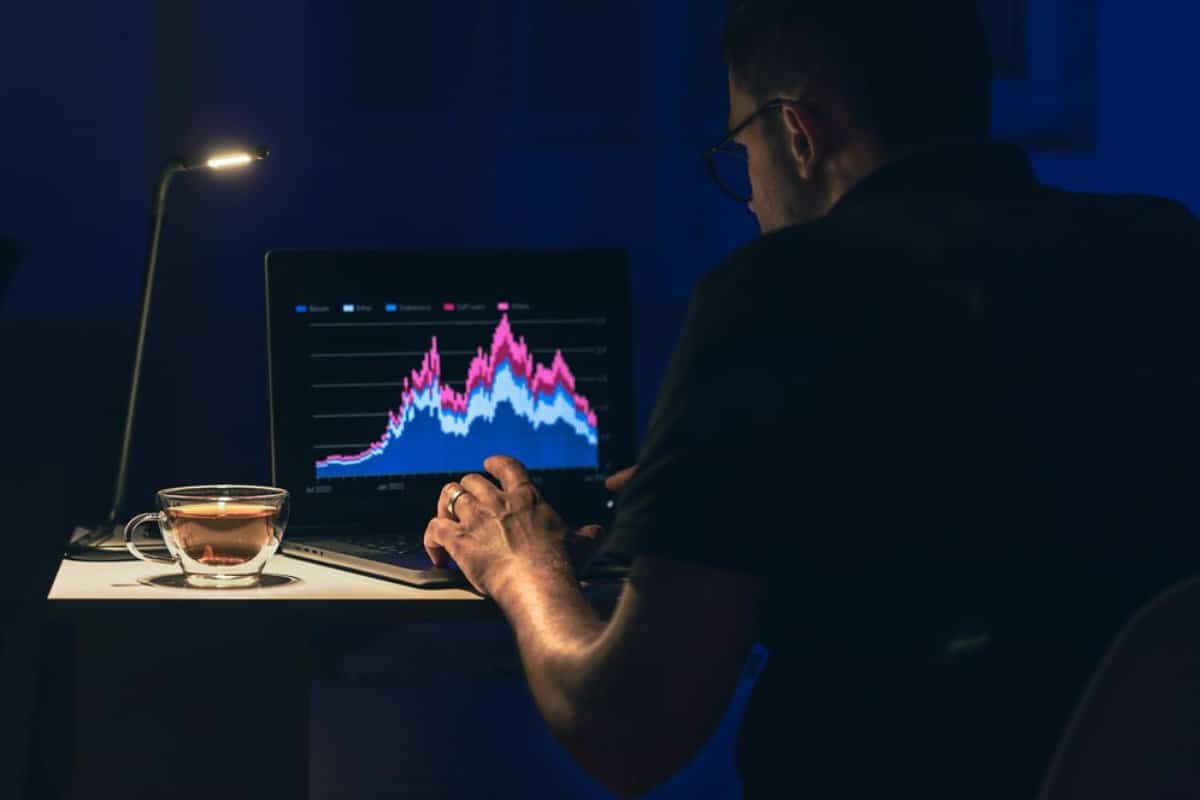 A man analyzes financial graphs on a laptop in a dimly lit room.